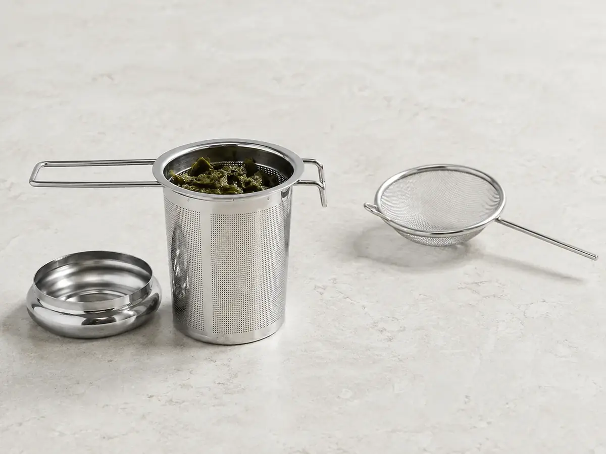 A stainless steel tea infuser basket holding wet leaves beside a small empty wire mesh hand strainer on a pale counter