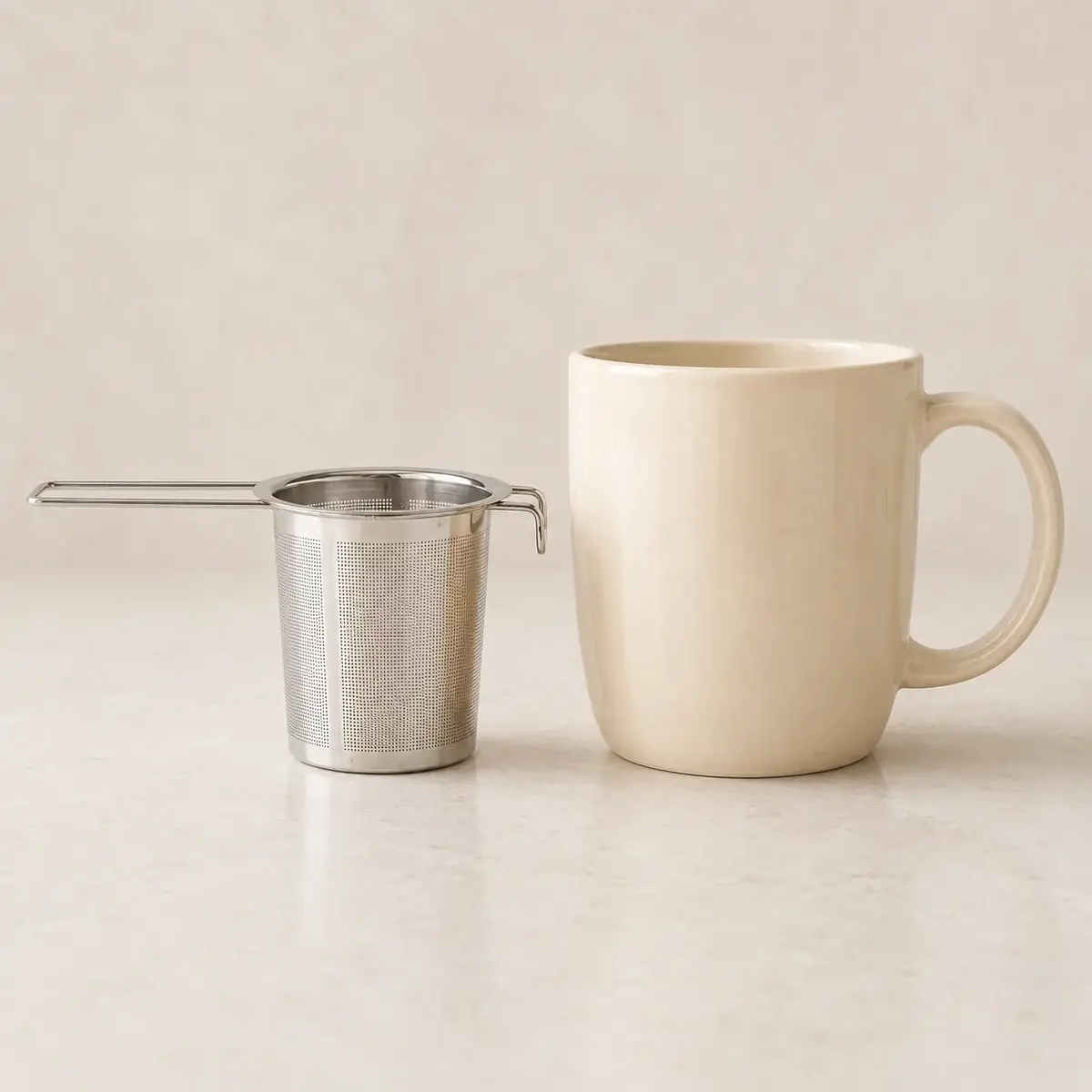 A steel infuser basket standing beside a plain ceramic mug on a pale counter, showing their relative size